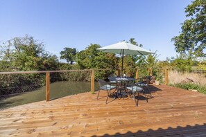 A wooden deck with a table and chairs, a white umbrella, and a view of a river.