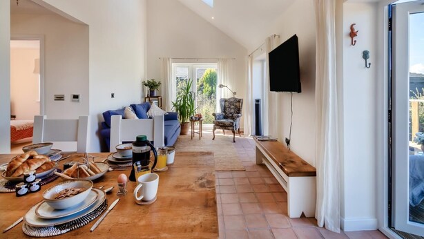 A spacious living area with a dining table set for breakfast, a sofa, a chair, and a TV mounted on the wall.