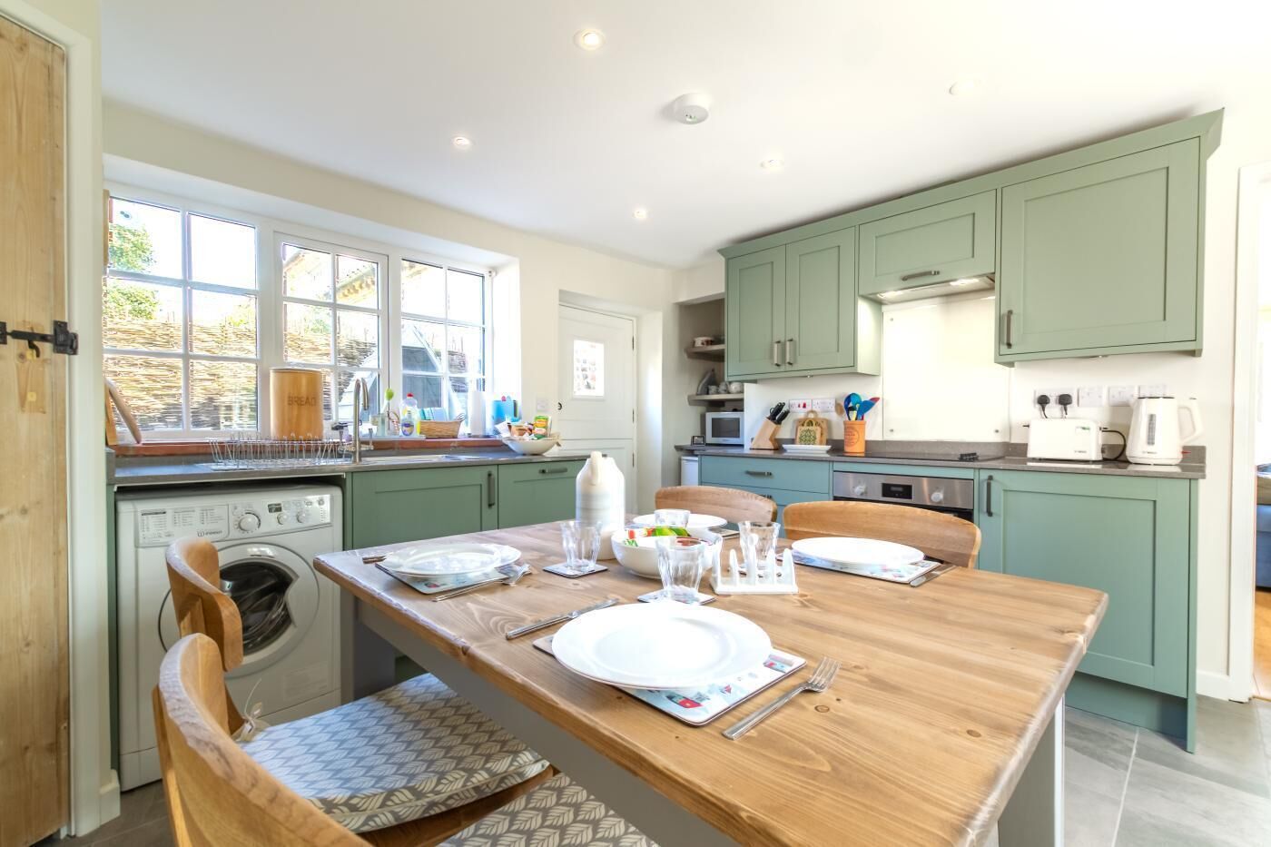 A kitchen with a wooden table, chairs, a washing machine, and a window with a view of greenery.