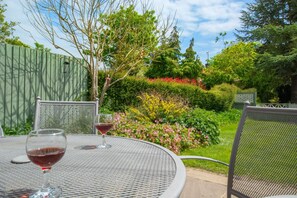 Una mesa metálica redonda con dos sillas, una copa de vino tinto, y un jardín con diversas plantas y una valla de madera al fondo.