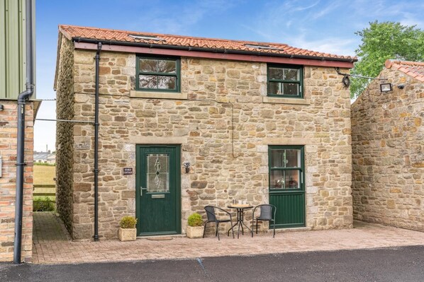 A stone building with a green door and windows, a small outdoor seating area with chairs, and a sign indicating a stable cottage. - 2 Bed in Crook (oc-s32614) (Crook)