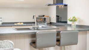 A modern kitchen with a built-in microwave, a stainless steel sink, and a black range hood. There are two grey bar stools and a vase with flowers on the counter. - 2 Bed in Crook (oc-s32614) (Crook)