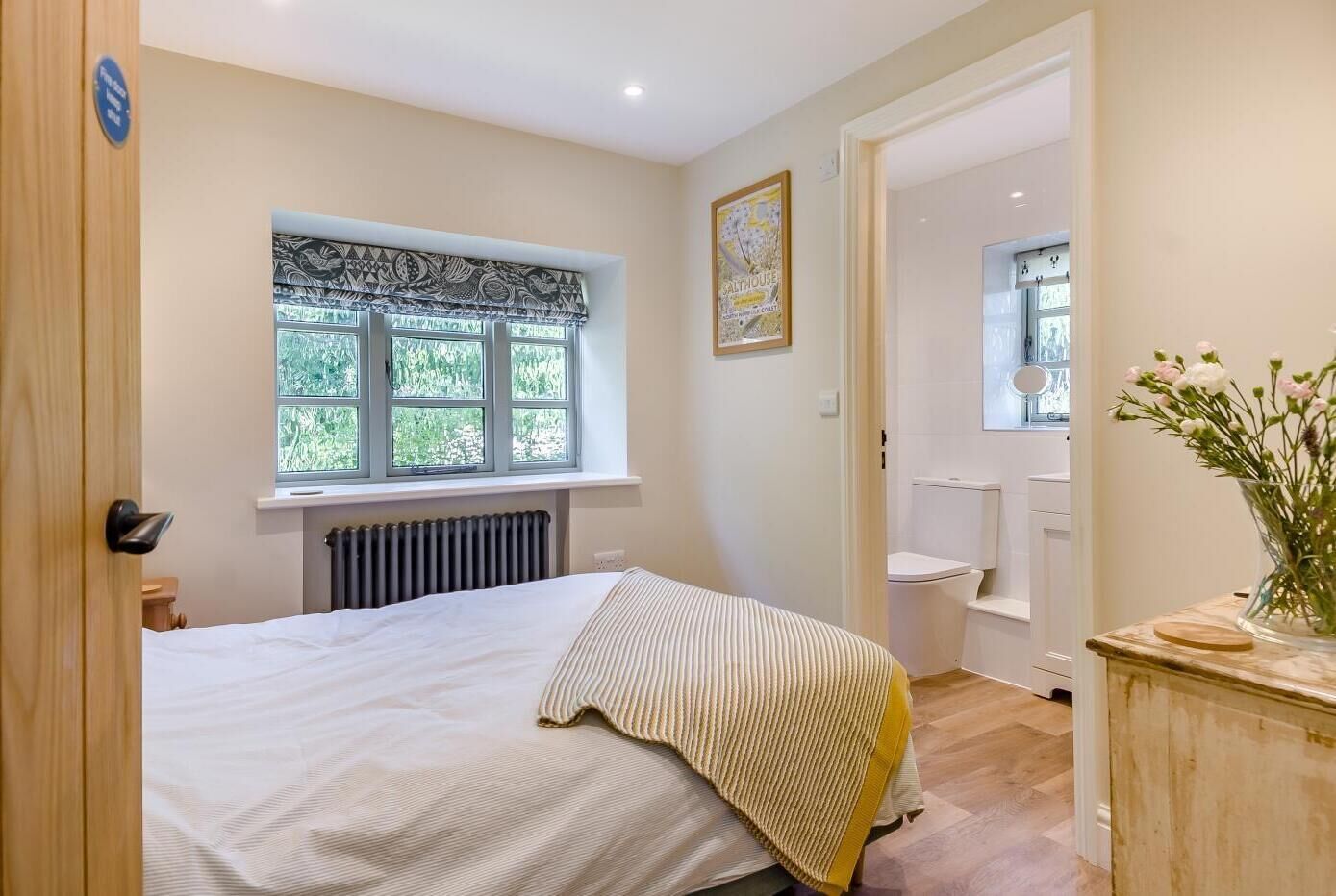 A bedroom with a bed, a radiator, a window with a view, a bathroom with a toilet, and a vase with flowers on a dresser.