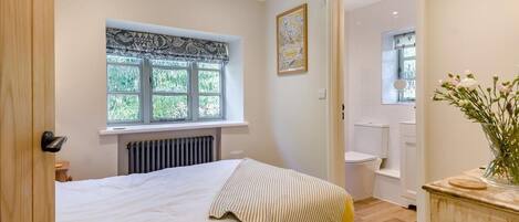 A bedroom with a bed, a radiator, a window with a view, a bathroom with a toilet, and a vase with flowers on a dresser.