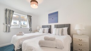 A hotel room with two beds, a window with curtains, a painting, and a bedside table with a lamp. - 2 Bed in Porthleven (oc-p00972) (Porthleven)