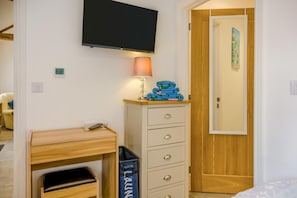 A bedroom with a bed, a desk with a lamp, a TV, a wooden door, and a trash bin. - 1 Bed in Gilsland (oc-h32687) (Waterhead)