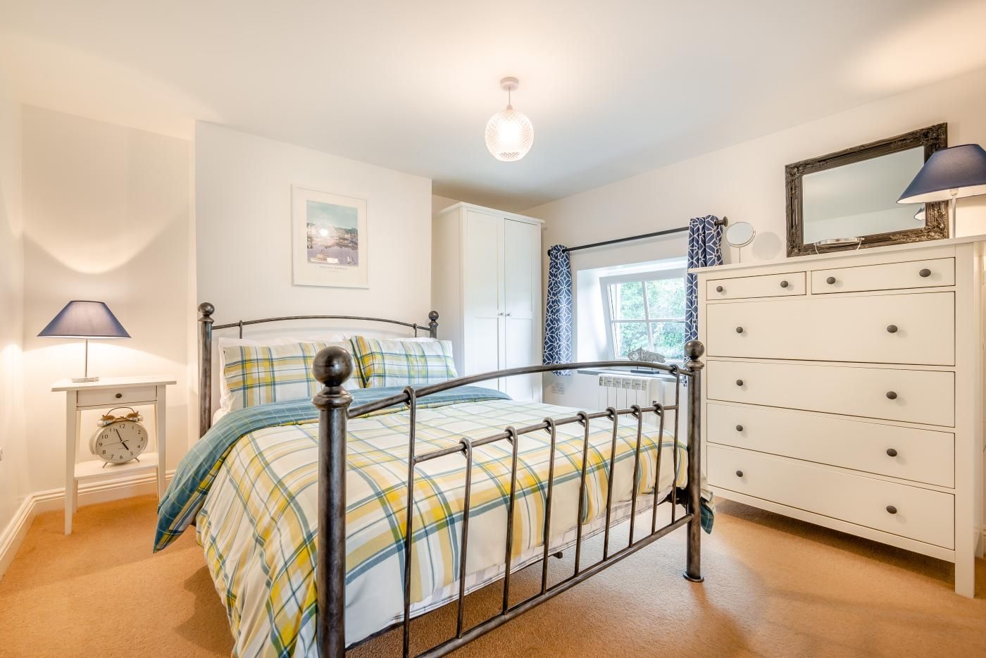 A bedroom with a metal bed frame, a white dresser, a bedside lamp, and a window with curtains.