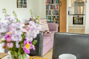 A living room with a sofa, a bookshelf, and a kitchen area with an oven. - 3 Bed in Padstow (oc-p00294) (Saint Issey)