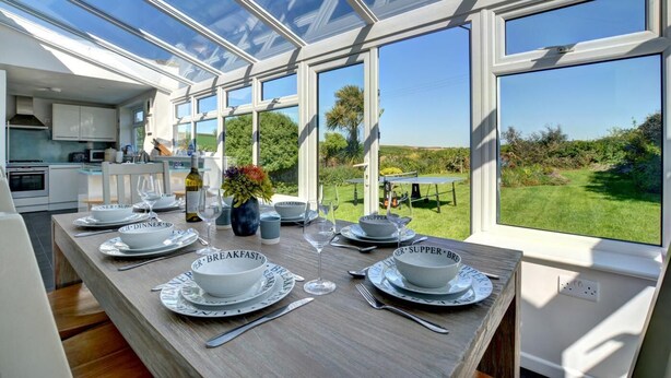 Ein Essbereich mit Tisch, gedeckt für Frühstück und Abendessen, großen Fenstern und Blick ins Freie.