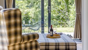 En mysig sittgrupp med en rutig soffa, ett träbricka med en flaska och ett glas, och utsikt över en uteservering med stolar och ett bord.