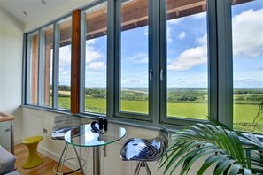 A room with large windows, a glass table, a yellow stool, and a potted plant. - 3 Bed in Wadebridge (oc-p00415) (Saint Breock)