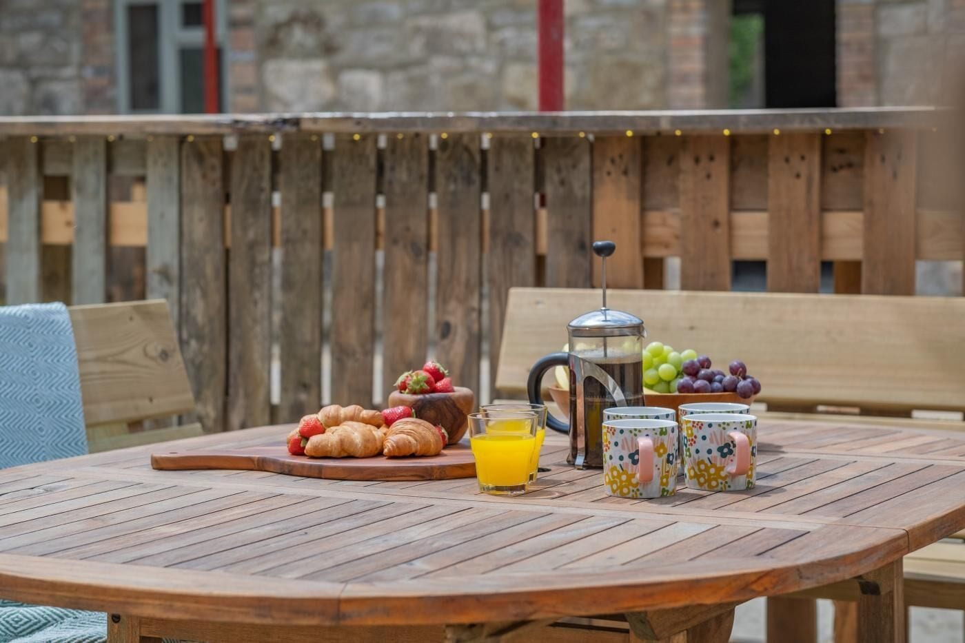 A wooden table with croissants, a fruit bowl, a coffee pot, and colorful mugs.