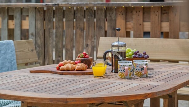 Ein Holztisch mit Croissants, einer Obstschale, einer Kaffeekanne und bunten Tassen.
