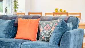 Ein blaues Sofa mit zwei Kissen, eines orange und eines mit einem Blumenmuster, in einem Raum mit einem Esstisch und Stühlen im Hintergrund.