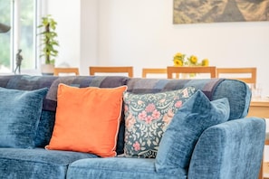 A blue sofa with two pillows, one orange and one with a floral pattern, in a room with a dining table and chairs in the background. - 3 Bed in Ventnor (oc-o32663) (Chale)