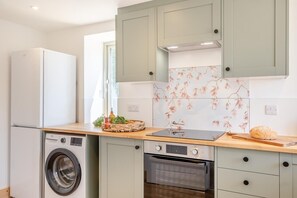 A compact kitchen with a washing machine, oven, and a wooden countertop with a basket of vegetables. - 3 Bed in Ventnor (oc-o32663) (Chale)
