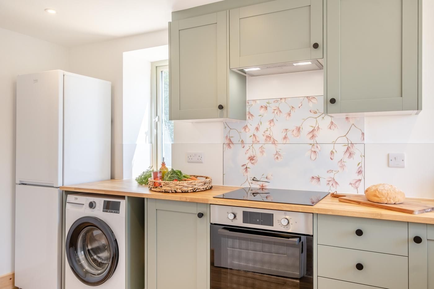 A compact kitchen with a washing machine, oven, and a wooden countertop with a basket of vegetables.