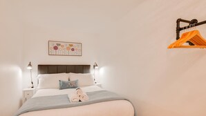 Una cama bien hecha con almohadas, una mesita de noche con una lámpara y un perchero en la pared con ropa.