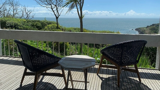 Una terraza de madera con dos sillas de mimbre y una mesita, con vistas a la costa, árboles y el océano.