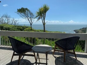 Eine Holzterrasse mit zwei Korbstühlen und einem kleinen Tisch, mit Blick auf die Küste, Bäume und das Meer.