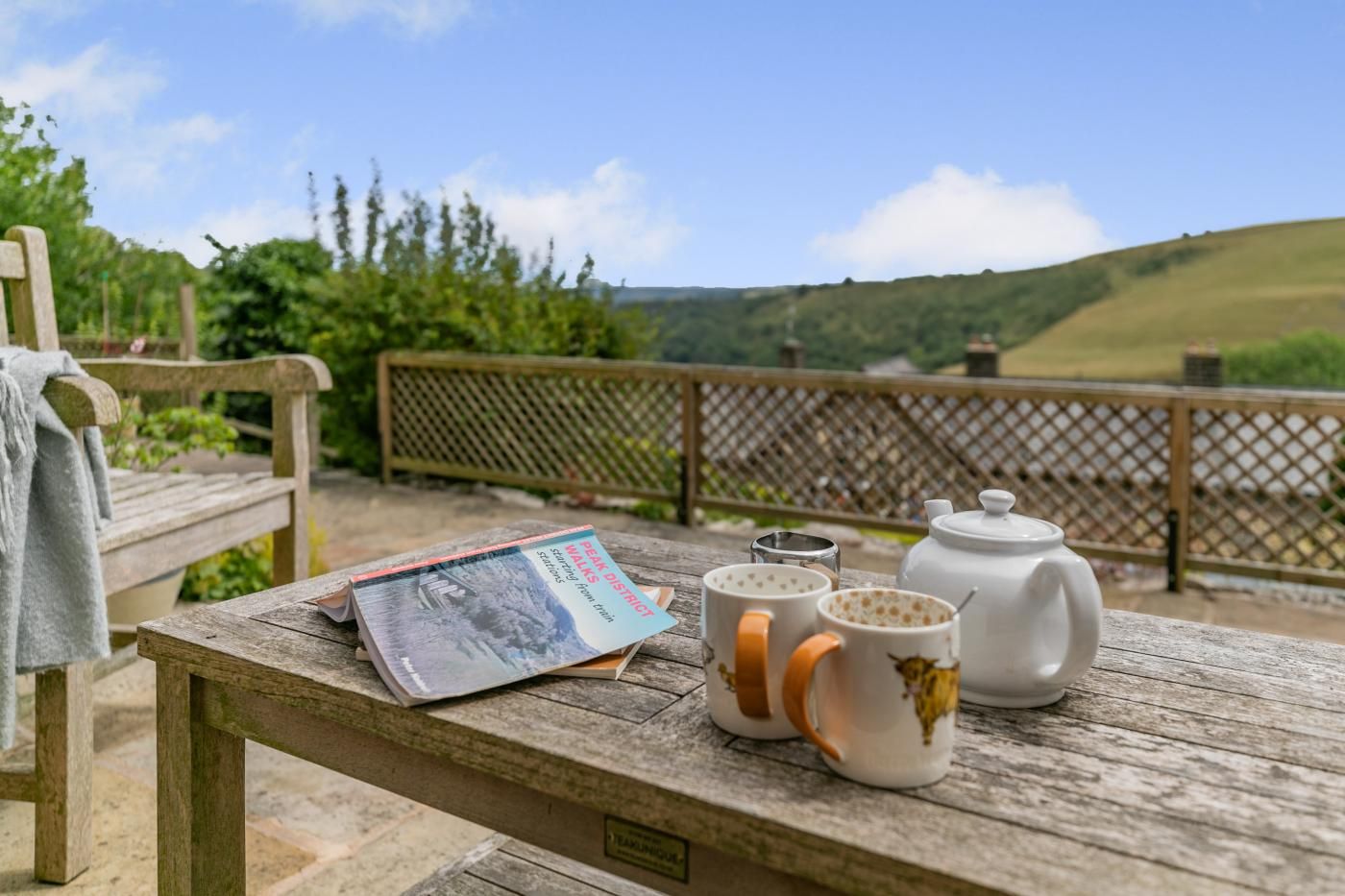 A wooden table with a book, two mugs, and a teapot on a balcony overlooking a green landscape.