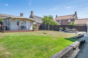 Il s’agit d’une propriété résidentielle dotée d’un jardin arrière enherbé, d’une terrasse en bois, d’un petit patio avec une table et des chaises, et d’une maison en briques surmontée d’un toit en tuiles.