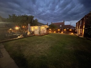 Un espace extérieur bien éclairé, équipé d’un barbecue, un petit bâtiment avec une horloge et, au fond, une maison en briques.