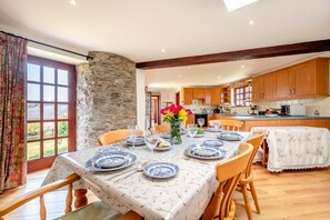Un coin repas avec une table dressée pour six personnes, entouré de chaises en bois, d’un mur en pierre et offrant une vue sur le jardin par le biais d’une grande fenêtre.