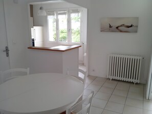 A white dining area with a round table and chairs, a small kitchen counter, a radiator, and a window with curtains.