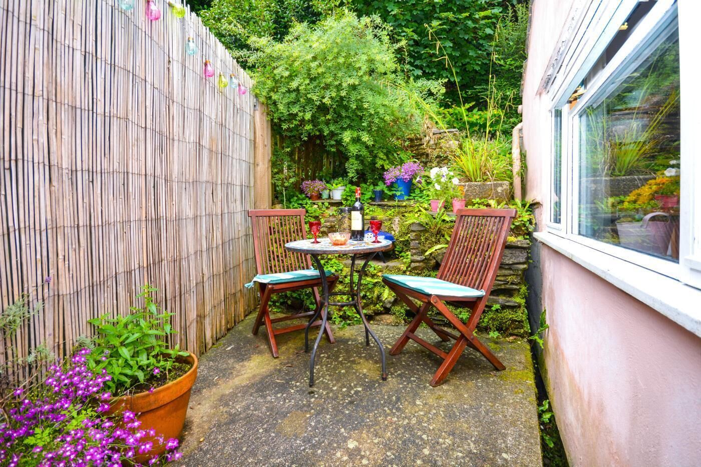 Une petite terrasse avec deux chaises en bois et une table, entourée d’une clôture en bambou et de diverses plantes.
