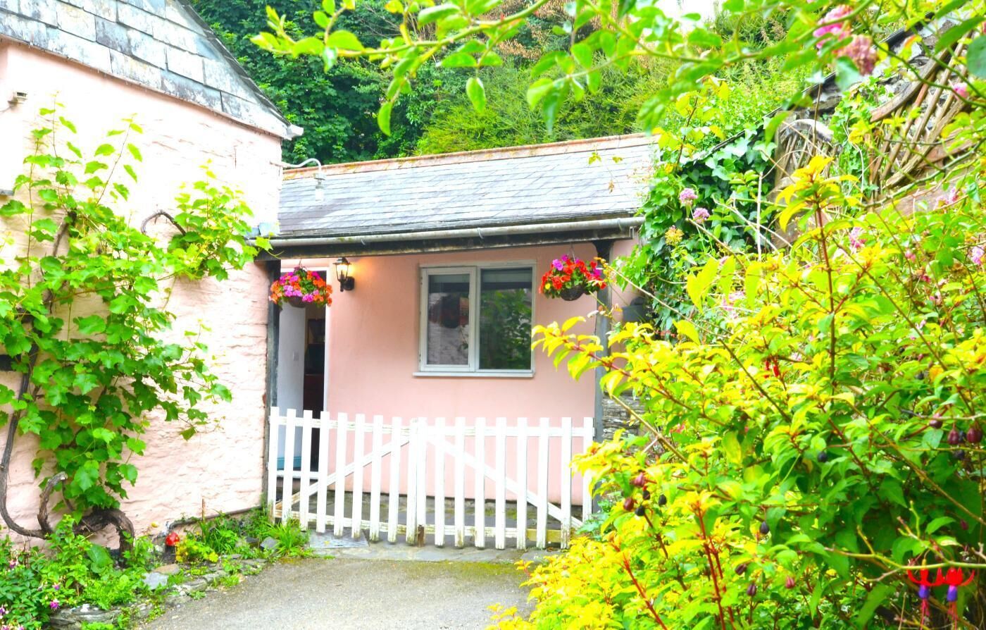 A small pink building with a white picket fence, surrounded by lush greenery and flowering plants.