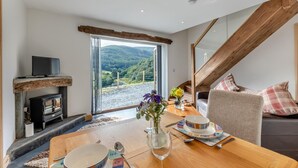 A modern living room with a stone fireplace, a dining table set with a vase of flowers, and a view of a mountainous landscape through large windows. - 1 Bed in Abergynolwyn (oc-p32345) (Abergynolwyn)
