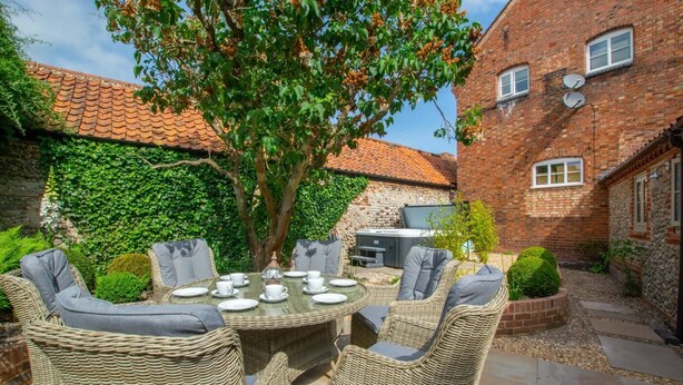 A patio with wicker furniture and a table set for dining, surrounded by greenery and a brick building. - 3 Bed in Holt (oc-2150) (Holt)