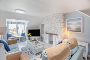 A modern living room with a stone fireplace, a flat-screen TV, and a large window overlooking a garden.