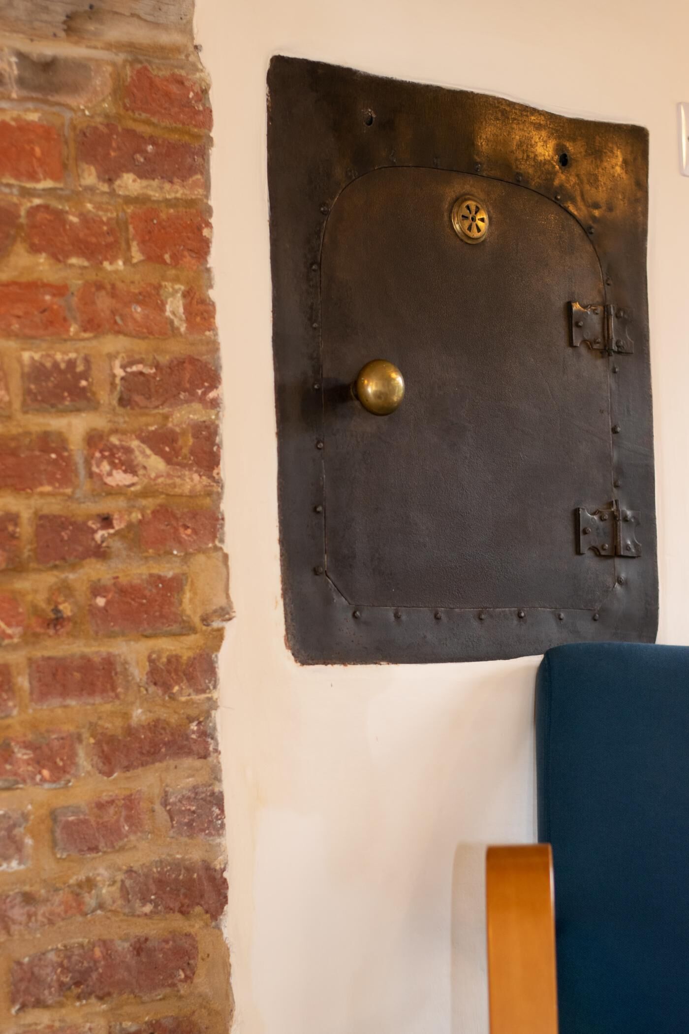 A wall-mounted door with a brass knob and a circular lock, adjacent to a brick wall and a blue upholstered piece of furniture.