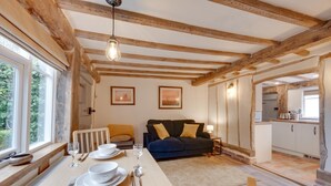 Ein gemütliches Zimmer mit Holzbalkendecke, einem Esstisch gedeckt mit Tellern, Gläsern und Besteck, einem Sofa mit Kissen und Blick auf eine Küchenzeile.