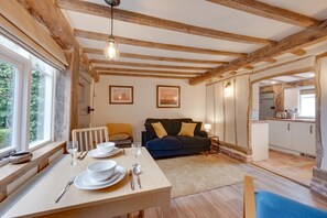 Ein gemütliches Zimmer mit Holzbalkendecke, einem Esstisch gedeckt mit Tellern, Gläsern und Besteck, einem Sofa mit Kissen und Blick auf eine Küchenzeile.