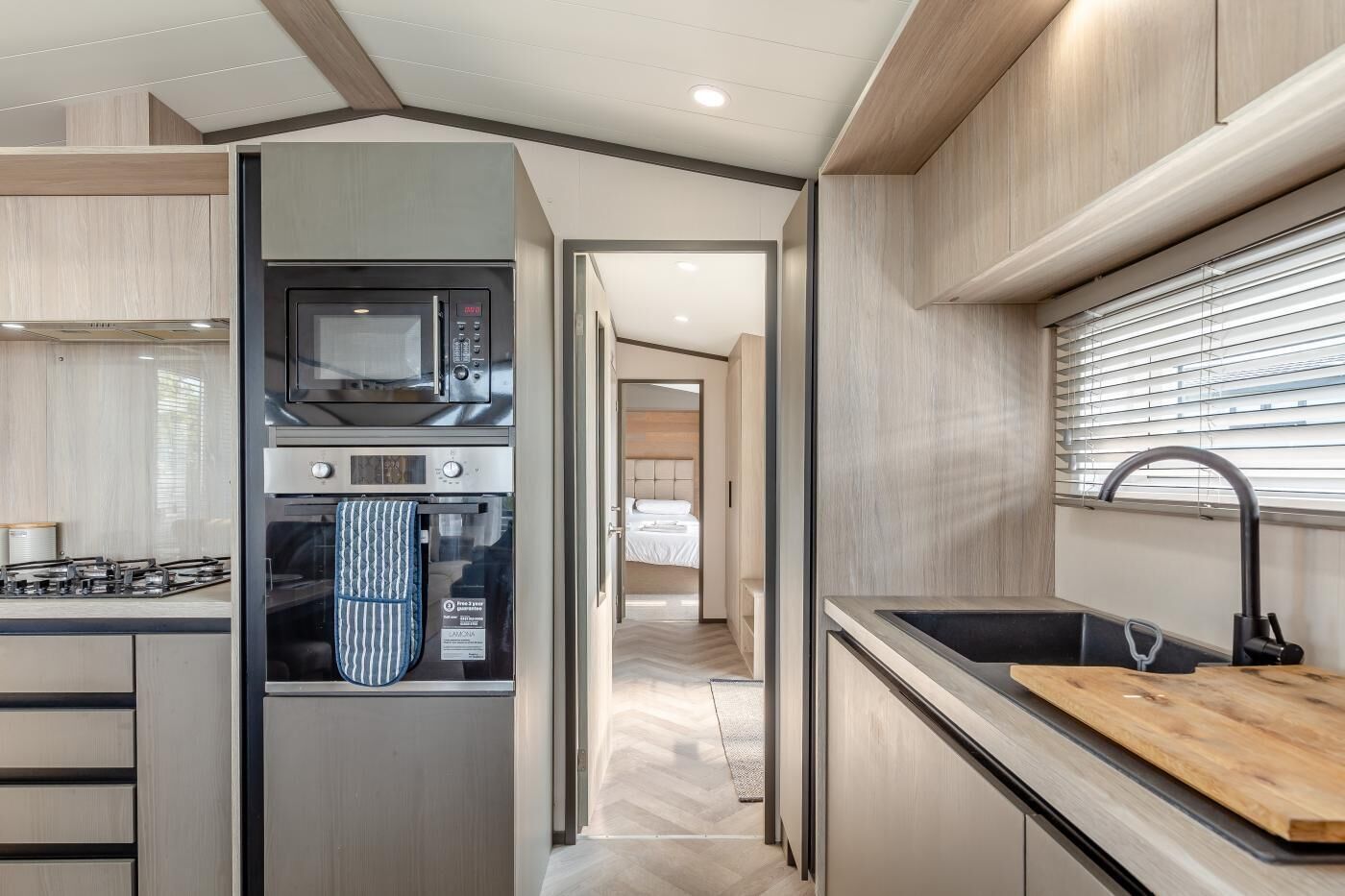 A compact kitchen with a microwave, oven, and gas stove. A wooden cutting board is on the counter, and a sink with a faucet is visible.