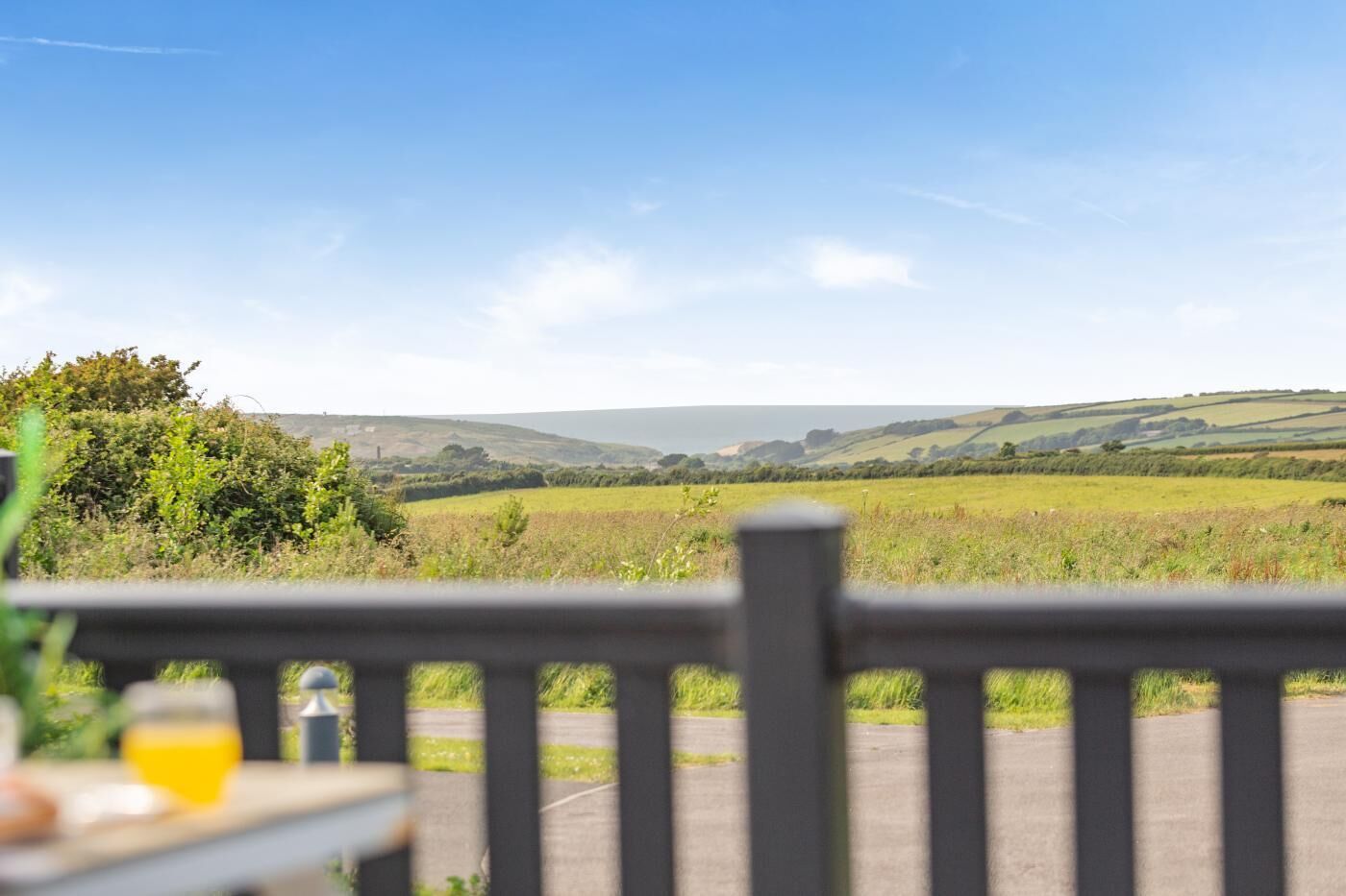 Une vue depuis un balcon, avec une table et un verre de jus de fruits, donnant sur un paysage rural avec des champs et des collines.