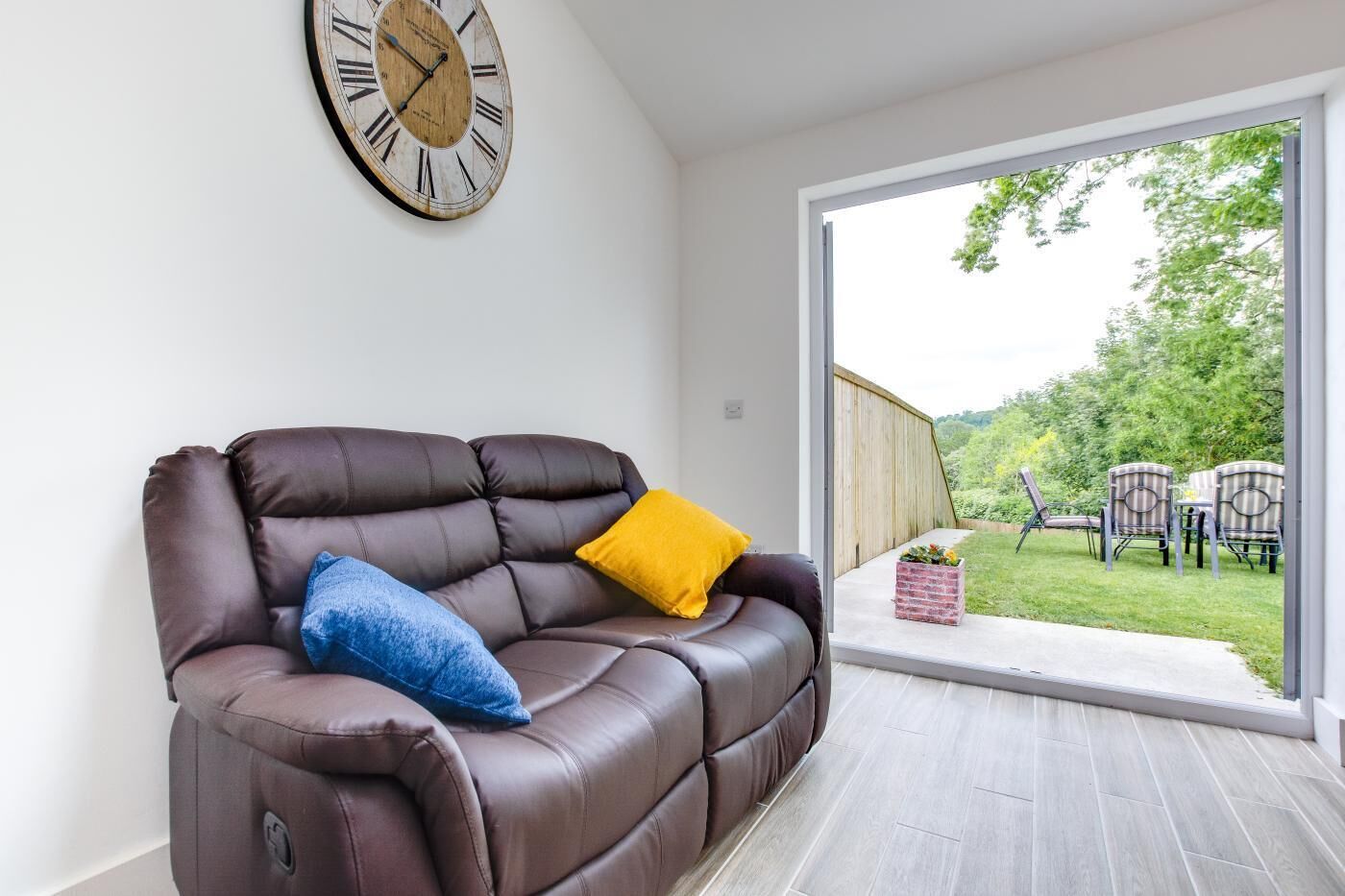 A modern living room with a leather sofa, a wooden clock, and a sliding glass door leading to an outdoor area with chairs and a garden.