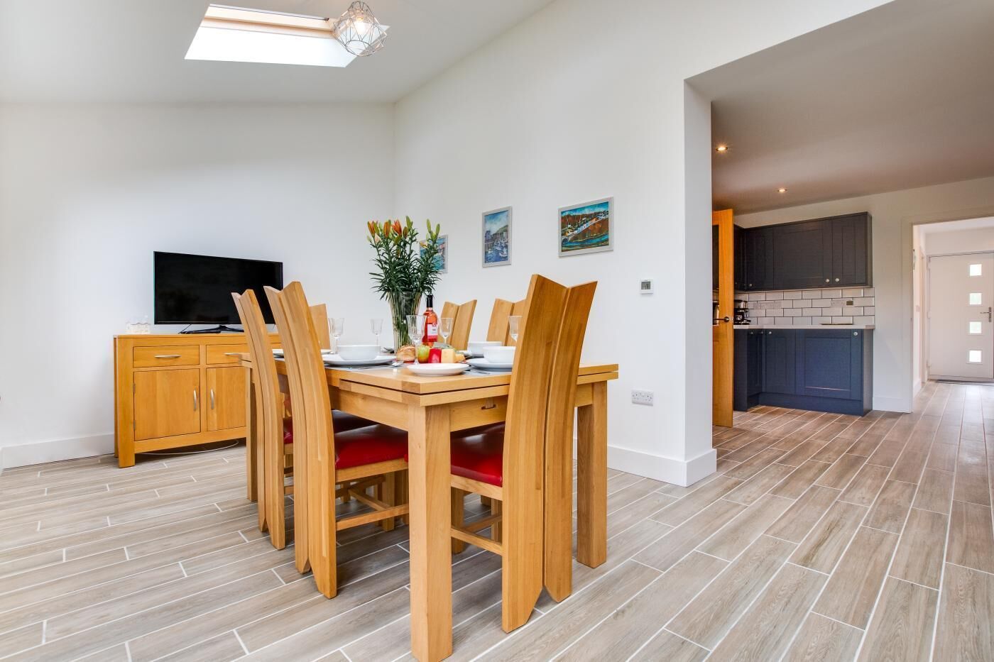 A dining area with a wooden table and chairs, a TV on a cabinet, and a kitchen in the background.
