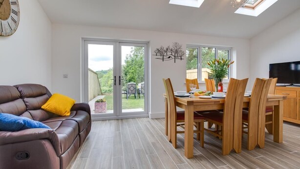A modern living room with a leather sofa, dining table, and chairs. There is a large window with a view of the outdoors. - 3 Bed in Llanfynydd (oc-pw9215) (Llanfynydd)
