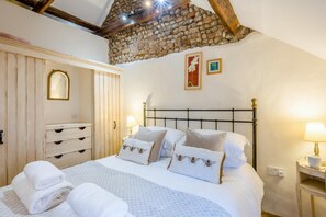 A bedroom with a stone ceiling, a bed with white bedding and bee-patterned pillows, a wooden wardrobe, and a nightstand with a lamp.