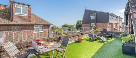 Un patio trasero residencial con césped verde, zona de estar exterior con sillas y una mesa, y una casa de ladrillo con chimenea.