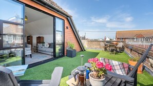 Ein modernes Haus mit Glastür, die zu einem Wohnzimmer führt, einer kleinen Sitzgelegenheit im Freien mit Tisch und Stühlen sowie einem Garten mit Rasen.