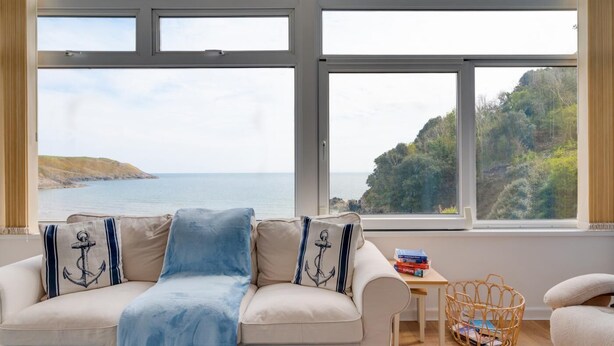 Habitación con un ventanal grande que ofrece vistas al mar y a las colinas. Hay un sofá con una manta azul y almohadas, una mesita con libros y una cesta de mimbre.