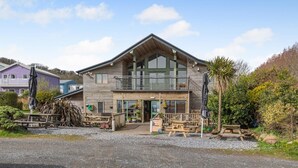 Un bâtiment en bois à deux étages, doté d’un balcon, d’un espace extérieur aménagé pour s’asseoir et d’une allée gravillonnée.