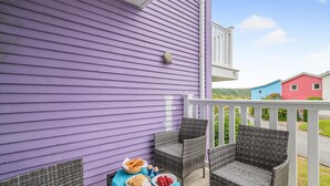 Un balcon agrémenté de fauteuils en osier et d’une table où sont disposés des plats, donnant sur une rue bordée de maisons aux couleurs vives.