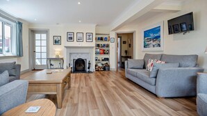 A spacious living room with a fireplace, grey sofas, wooden flooring, and a bookshelf. - 7 Bed in Aldeburgh (oc-midba) (Aldeburgh)