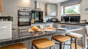 A modern kitchen with built-in appliances, a large island with stools, and a window with a view of greenery. - 7 Bed in Aldeburgh (oc-midba) (Aldeburgh)
